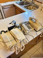 Overview of four telephones on counter: beige desk phone, two beige wall-mount phones, black cordless handset with base and cords