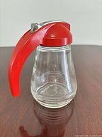 Full view of glass syrup dispenser with red plastic handle and metal lever