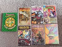 Group shot of six TMNT comic books and one TMNT Pizza Cookbook arranged on carpet