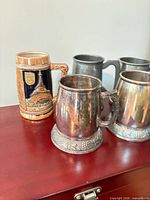 Group view showing ceramic Eiffel Tower stein, two silver-plated ornate tankards, two plain pewter tankards