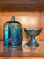Covered jar and footed compote side by side displaying iridescent finish and grape motif