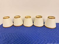Five ivory bell lampshades lined up upright showing exterior fabric and gold trim