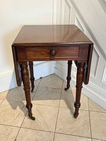 Front view showing drop leaves down, functional drawer with pull, turned legs, brass casters