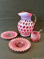 Fenton cranberry coin dot pitcher, hobnail tumbler with spoon, two hobnail candy dishes displayed on dark background