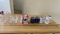 All nine mugs lined on tabletop including clear glass, boxed, speckled and printed ceramic pieces