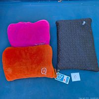 Three travel pillows/throw set on blue background: orange unit with tags, pink pillow, black quilted inflatable pillow