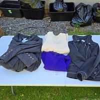 Four garments folded on table: grey coat, yellow sweater, purple sweater, black TanJay jacket
