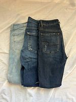 Three pairs of jeans stacked showing back pockets, light to dark washes