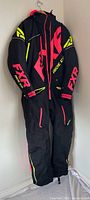 Front view of black FXR one-piece snow suit hung on wall, showing pink logos, yellow accents, hood, full-length zipper.