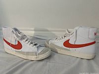 Pair of white high-top Nike Blazer Mid ’77 Vintage sneakers with orange swooshes displayed side by side