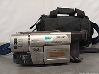Left side of camcorder showing model, zoom rating, attached strap and carrying case in background
