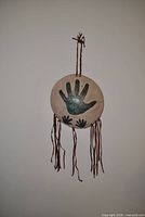 Front view of circular beige ceramic plaque with green handprint and leather fringe