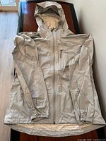 Front view of light gray hooded rain jacket laid flat showing zip closure and pockets
