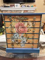 Front view of jewelry box showing six drawers, floral artwork, blue frame, gold knobs