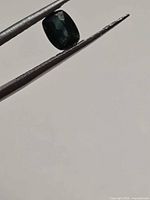 Cushion cut blue-green sapphire held by tweezers, angled view