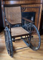 Front angled view of wheelchair showing wood frame, cane seat/back and metal rear wheels