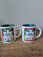 Front view of both mugs showing Mom and Dad text and holly border