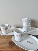 Four mugs and four plates displayed on tabletop showing logo and shapes