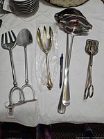 Group of large serving utensils including stirrup spatulas, serving spoon, gold-tone fork, ladle, scissor pastry server
