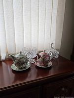 Two floral tea-for-one sets and three crystal dishes displayed on wooden surface