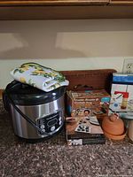 All items grouped on countertop: rice cooker, towel, boxed rosette iron set, boxed cookie press, terracotta garlic keeper, boxed letter appetizer dishes