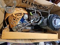 Box containing various kitchen items including blender jar, egg beater, glass measuring cup, wood tray, basket