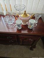 Group of crystal vase, two crystal bowls, pink teapot and two floral mugs on tabletop