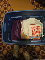Plastic bin with folded linens: purple knit piece, lace-trimmed white round cloth, red and white gingham napkins