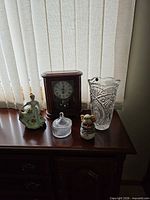 All five items on wooden sideboard: mantel clock, porcelain lady, lidded candy dish, teddy bear figurine, tall crystal vase