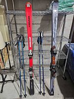 Two pairs skis and two pairs poles standing against shelving