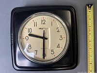 Front view of square black wall clock with measuring tape indicating 9 in width