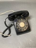 Black rotary dial telephone with handset, coiled handset cord, and line cord on neutral background