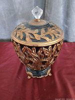 Full view of black and gold resin urn with ornate foliage relief and glass finial