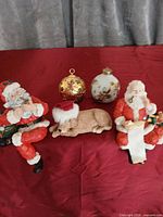 Group shot of two Santa figurines, dog figurine and two ornaments on red cloth