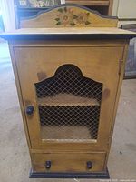 front view of distressed yellow cabinet showing wire mesh door, floral crest, drawer