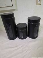 Three black canisters standing with lids on, size comparison