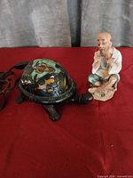 Turtle stained glass lamp and seated porcelain man figurine side by side on red cloth