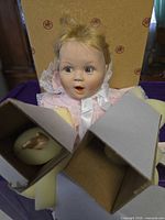 Porcelain baby doll in pink outfit inside original box with foam arm inserts visible