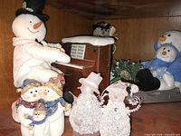 Shelf view showing ceramic piano snowman, trio figurine, glass snowmen, plush snowman in sled