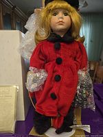 Front view of Blanche porcelain doll in red coat with bubble wrap on arms, standing on display stand