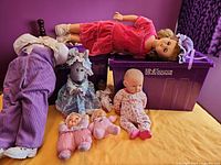 Group shot showing all seven dolls and plush on yellow surface with purple bins behind