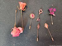 Six of the stick pins laid out: two resin roses, filigree oval, novelty key, purple pansy, shell cameo