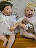 Both baby angel dolls seated side by side showing costumes, halos, wings and teddy bear accessory