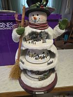 Front view of three-tier musical snowman showing village scenes, broom, scarf, mittens, and wood base