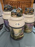 Three Thomas Kinkade ceramic canisters with resin cottage lids displayed together