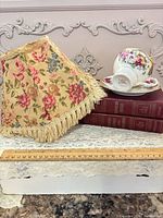 Floral fabric lampshade, stack of three red cloth-bound books, Castle China teacup and saucer with ruler for scale