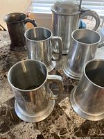 Five pewter mugs and one darker silver-plated mug on tabletop