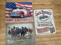 Front view of three printed metal signs showing Chevrolet pickup, Jack Daniel's whiskey ad, and horse welcome design