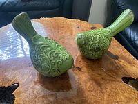 Front three-quarter view of both green ceramic bird figurines on wood surface