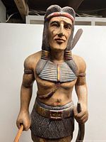 front view of carved wooden Indigenous warrior statue showing torso, bow, tomahawk and paint details